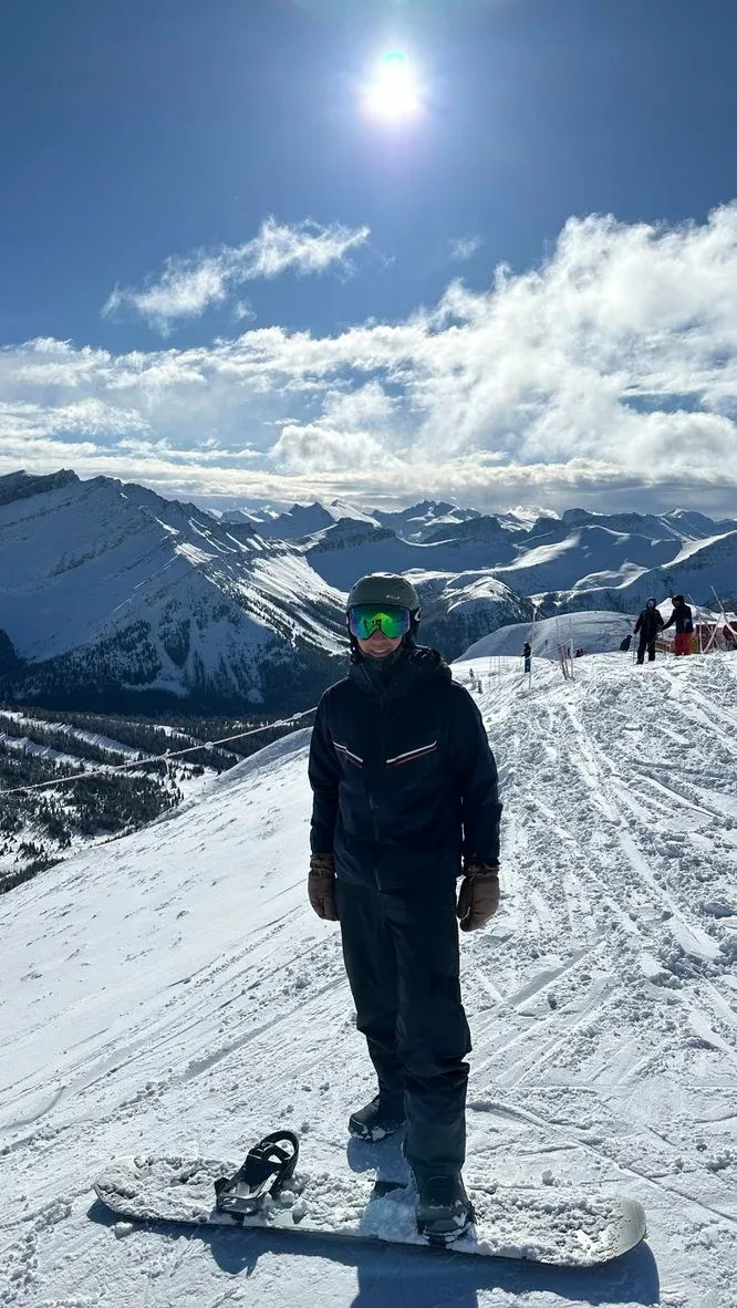 Andrew standing with his snowboard at the top of a run, snow-covered peaks behind him.